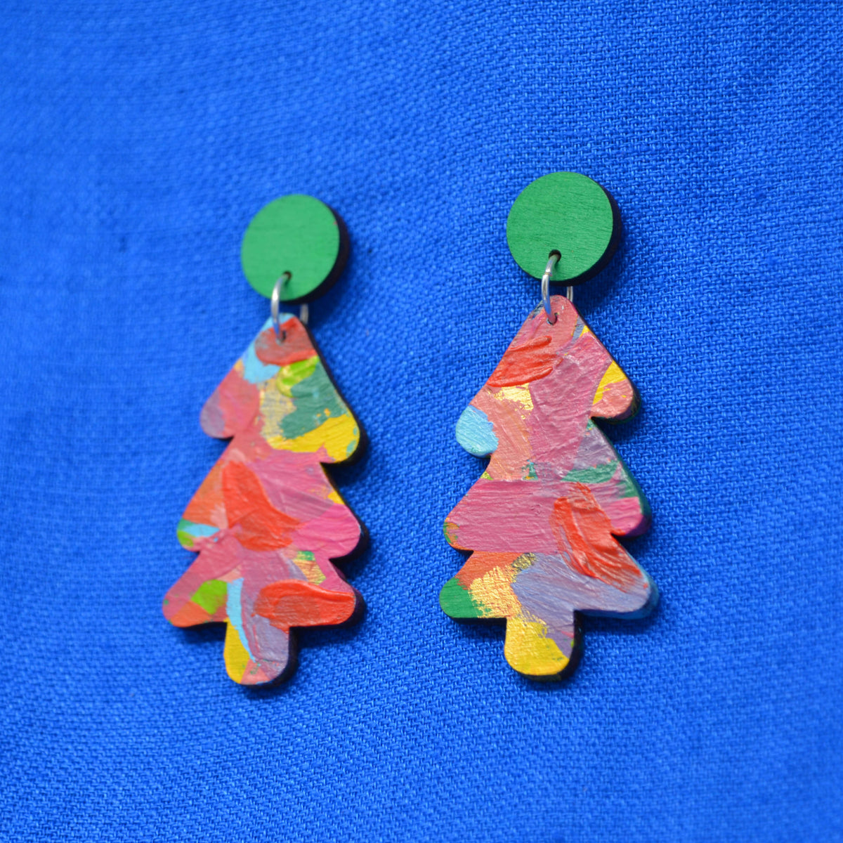 A pair of Christmas tree-shaped earrings with a colorful, textured paint-stroke pattern in pink, orange, yellow, and green, hanging from solid green circular studs against a bright blue textured fabric background.