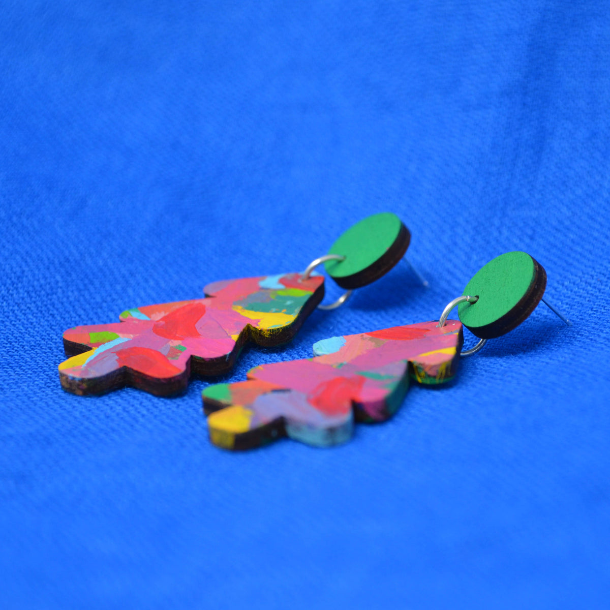 A pair of colorful tree-shaped wooden earrings with a multicolor abstract pattern and green circular studs, displayed on a textured blue background.