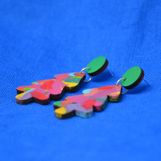 A pair of colorful tree-shaped wooden earrings with a multicolor abstract pattern and green circular studs, displayed on a textured blue background.