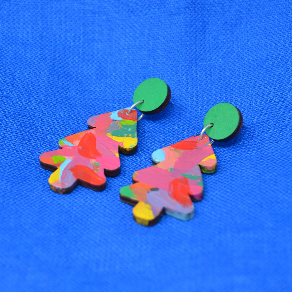 A pair of colorful, tree-shaped drop earrings with a marbled paint pattern in pink, red, yellow, and green, featuring solid green circular studs, displayed on a textured blue background.