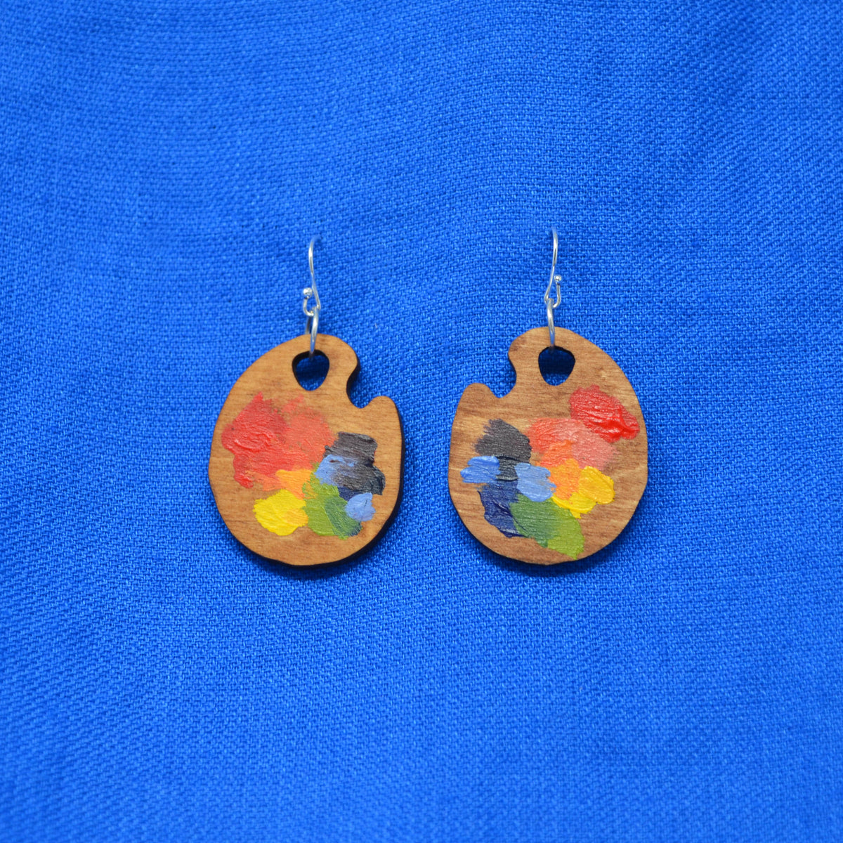 A pair of wooden painter's palette earrings with colorful paint details, hanging from silver fishhook wires against a bright blue textured fabric background.