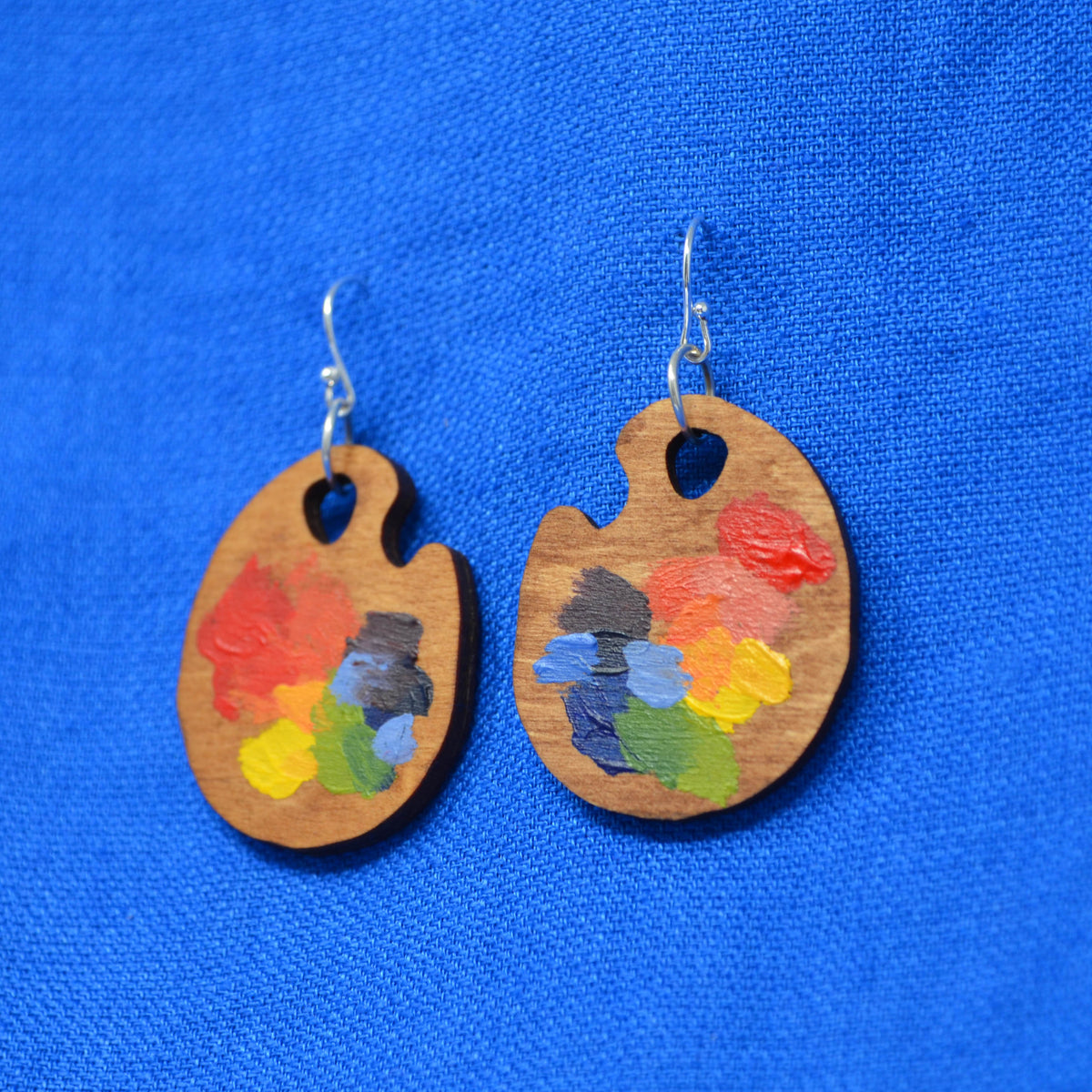 A pair of wooden artist paint palette earrings with colorful hand-painted paint strokes, shown on a textured bright blue background.