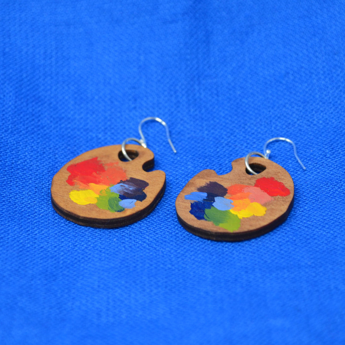 A pair of wooden painter's palette earrings with colorful paint swatches and silver fishhook wires, displayed on a textured blue background.