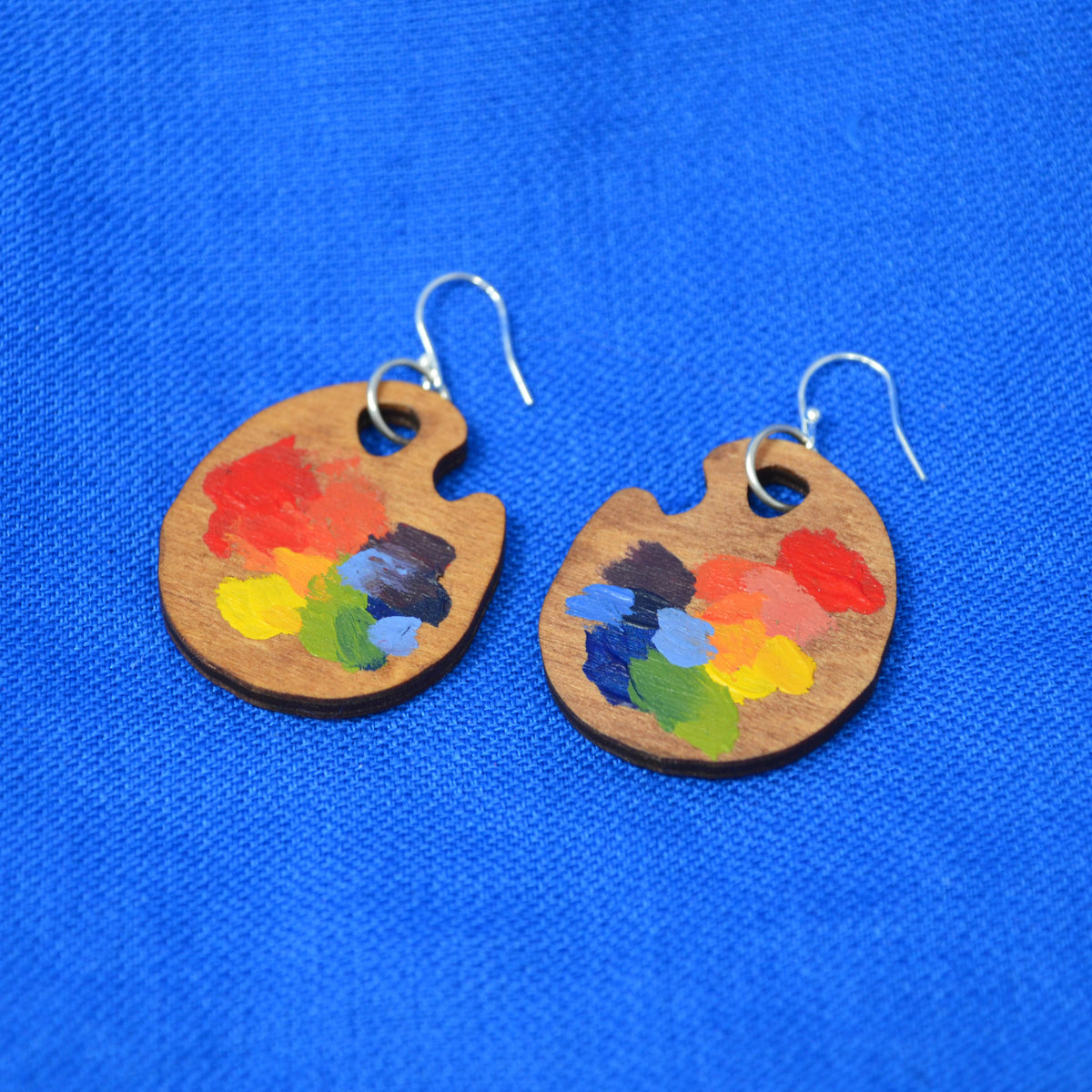 A pair of wooden artist palette earrings with silver fishhook wires, featuring painted multicolor paint strokes on a blue textured fabric background.