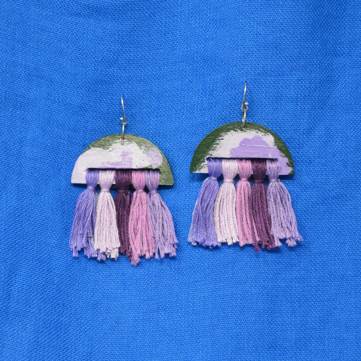 A pair of handmade dangle earrings featuring a semi-circular top piece with a green, purple, and white painted pattern, hanging from silver hooks, with multiple colorful tassels in shades of purple, pink, and white below. The earrings are displayed on a textured blue fabric background.