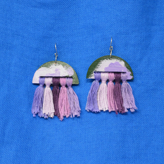 A pair of handmade dangle earrings featuring a semi-circular top piece with a green, purple, and white painted pattern, hanging from silver hooks, with multiple colorful tassels in shades of purple, pink, and white below. The earrings are displayed on a textured blue fabric background.