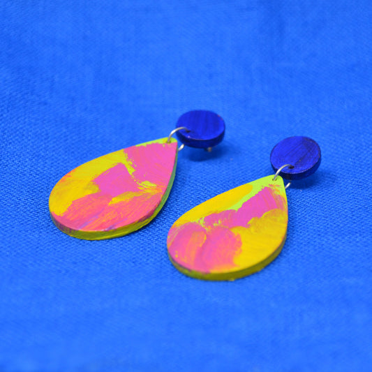 A pair of handmade wooden drop earrings with dark blue circular studs and teardrop shaped dangles featuring a pink, green and yellow abstract painted design, displayed on a textured blue background.