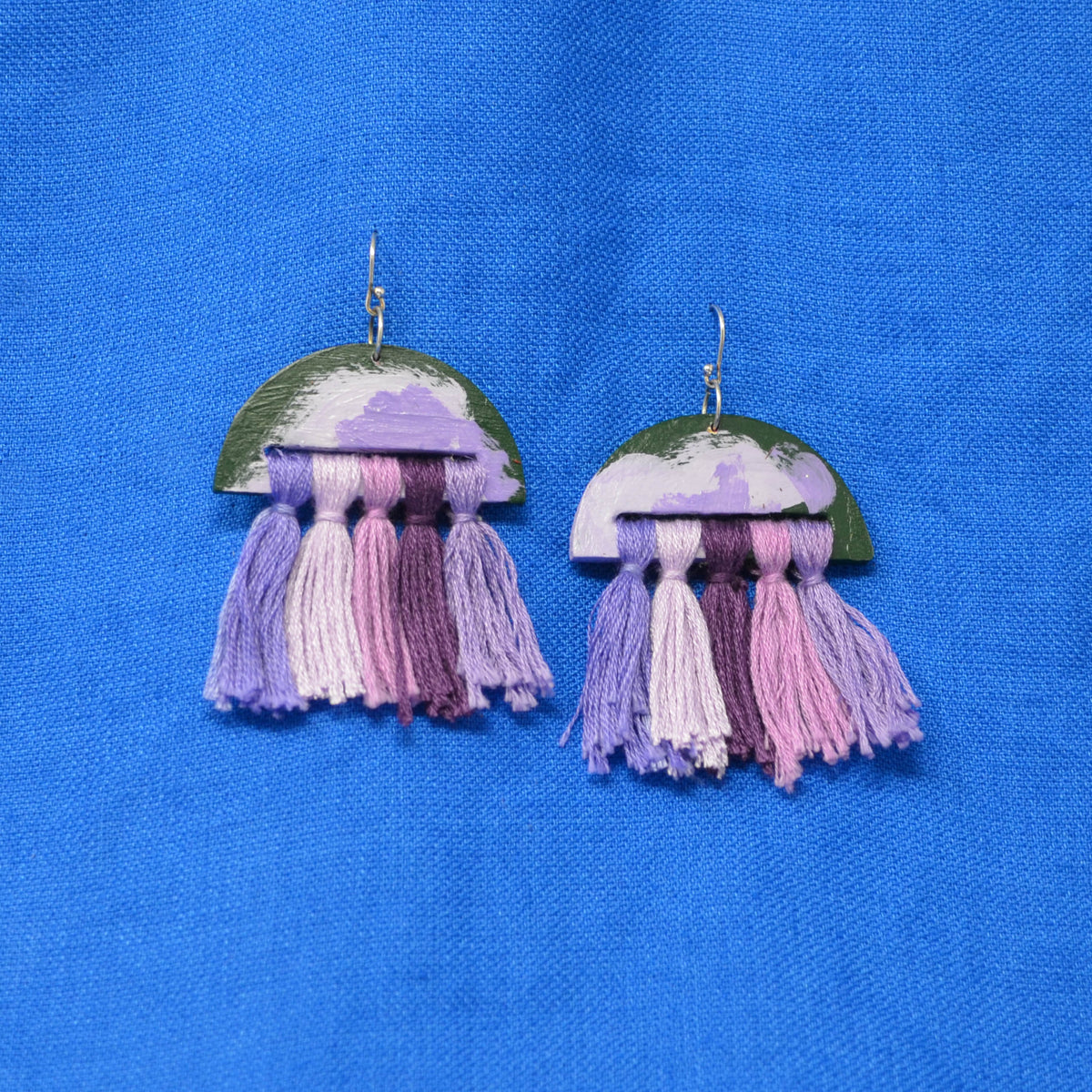 A pair of handmade jellyfish-inspired drop earrings with painted green and purple halos and dangling tassels in shades of lavender and purple, displayed on a textured blue fabric background.