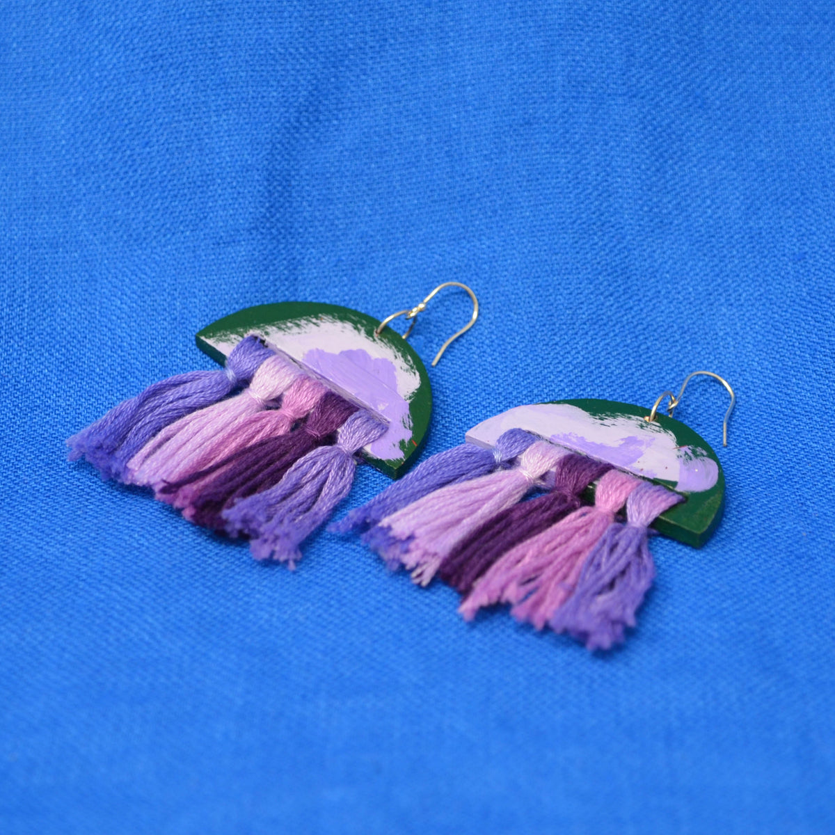 A pair of handmade jellyfish-inspired dangle earrings with purple and pink gradient tassels, green backs, and silver fishhook wires, displayed on a textured blue fabric background.