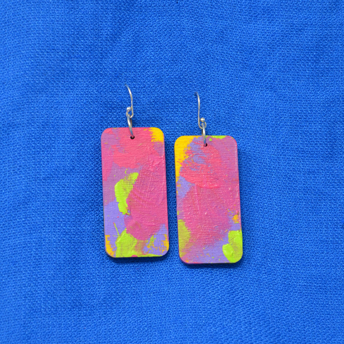 A pair of rectangular drop earrings with a vibrant, abstract paint-splatter design in pink, purple, yellow, and green, hanging from silver fishhook wires on a textured blue background.