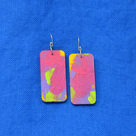 A pair of rectangular drop earrings with a vibrant, abstract paint-splatter design in pink, purple, yellow, and green, hanging from silver fishhook wires on a textured blue background.