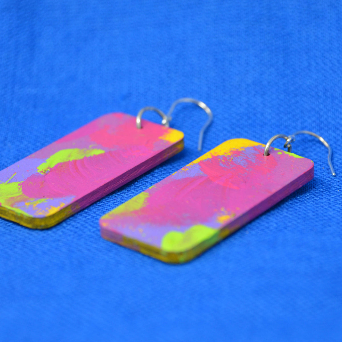 A pair of rectangular drop earrings with a vibrant pink, yellow, and green abstract paint design, attached to silver fishhook wires, displayed on a textured blue background.
