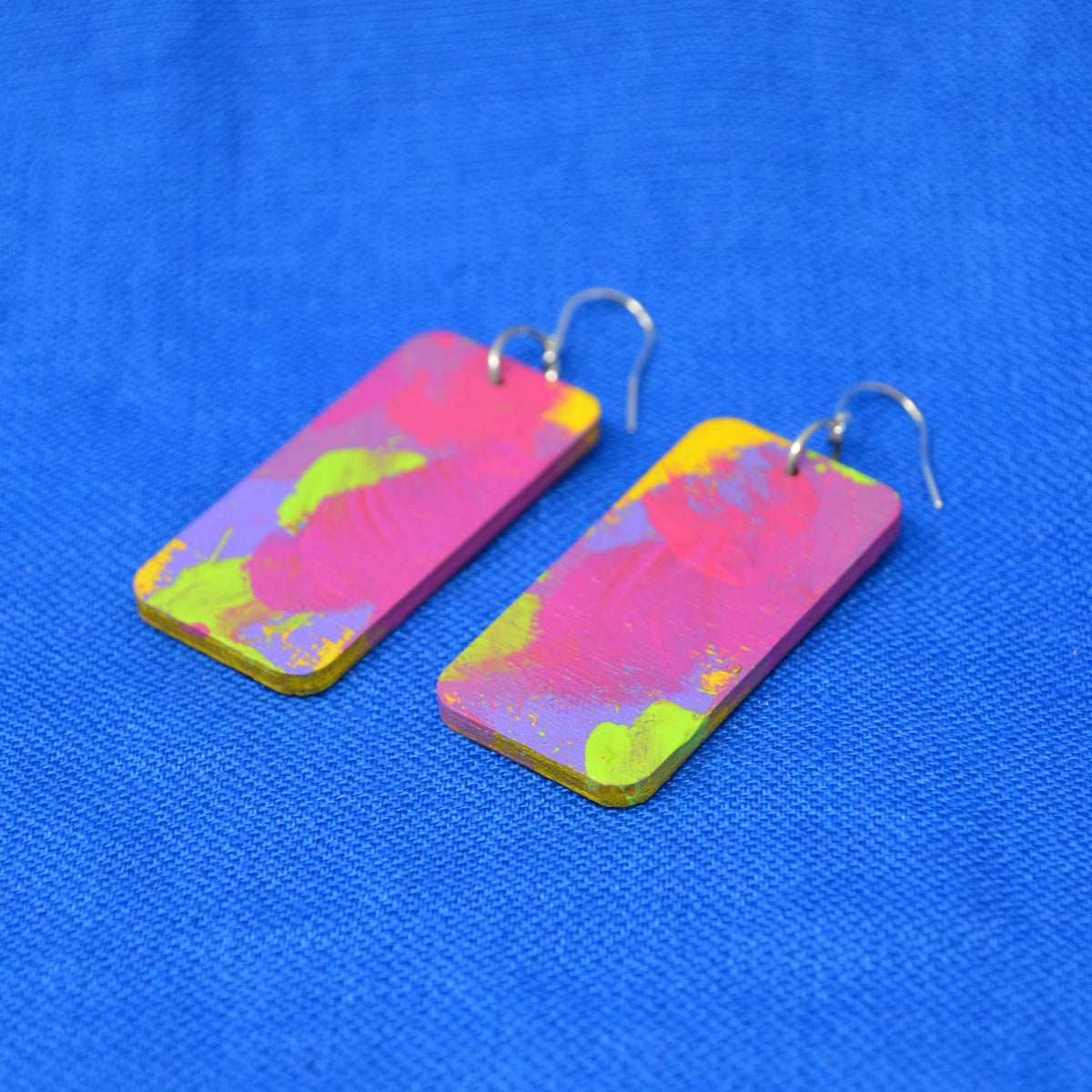 A pair of rectangular drop earrings with a vibrant pink, yellow, and green abstract paint design, attached to silver fishhook wires, displayed on a textured blue background.