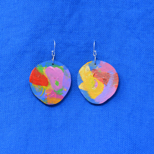 A pair of handmade oval drop earrings with colorful abstract painted designs in pink, yellow, orange, green, and blue, hanging from silver fishhook wires on a bright blue textured fabric background.