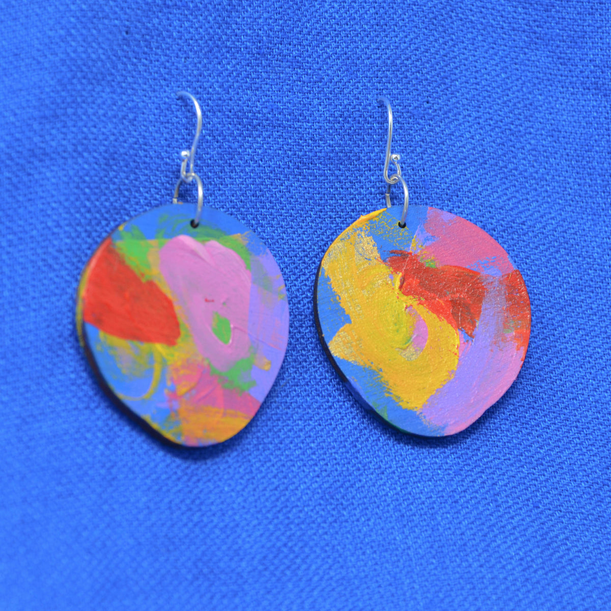 A pair of handmade earrings made from wood, featuring an abstract multicolor design with pink, yellow, blue, red, and green paint strokes, hanging from silver fishhook wires against a textured blue background.