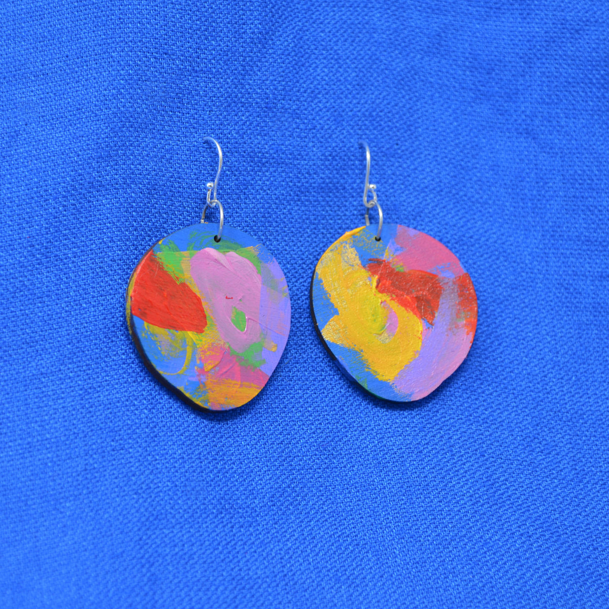 A pair of handmade drop earrings made from wood, painted with abstract multicolor brushstrokes, hanging from silver fishhook wires against a textured blue background.