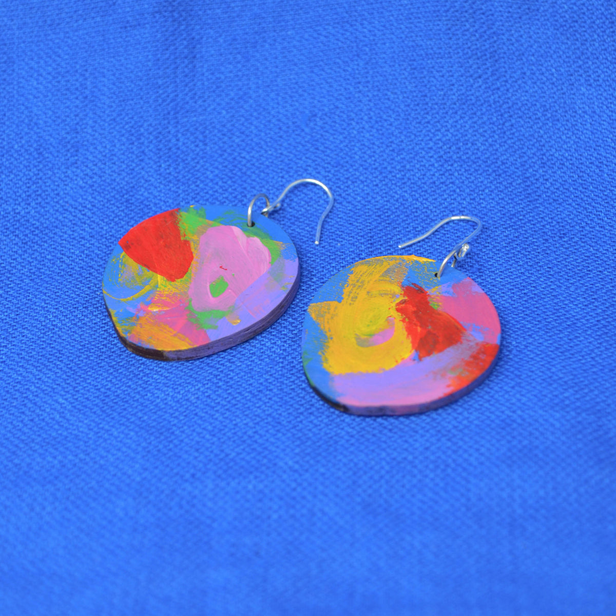 A pair of wooden drop earrings with abstract multicolor designs, displayed on a textured blue fabric background.