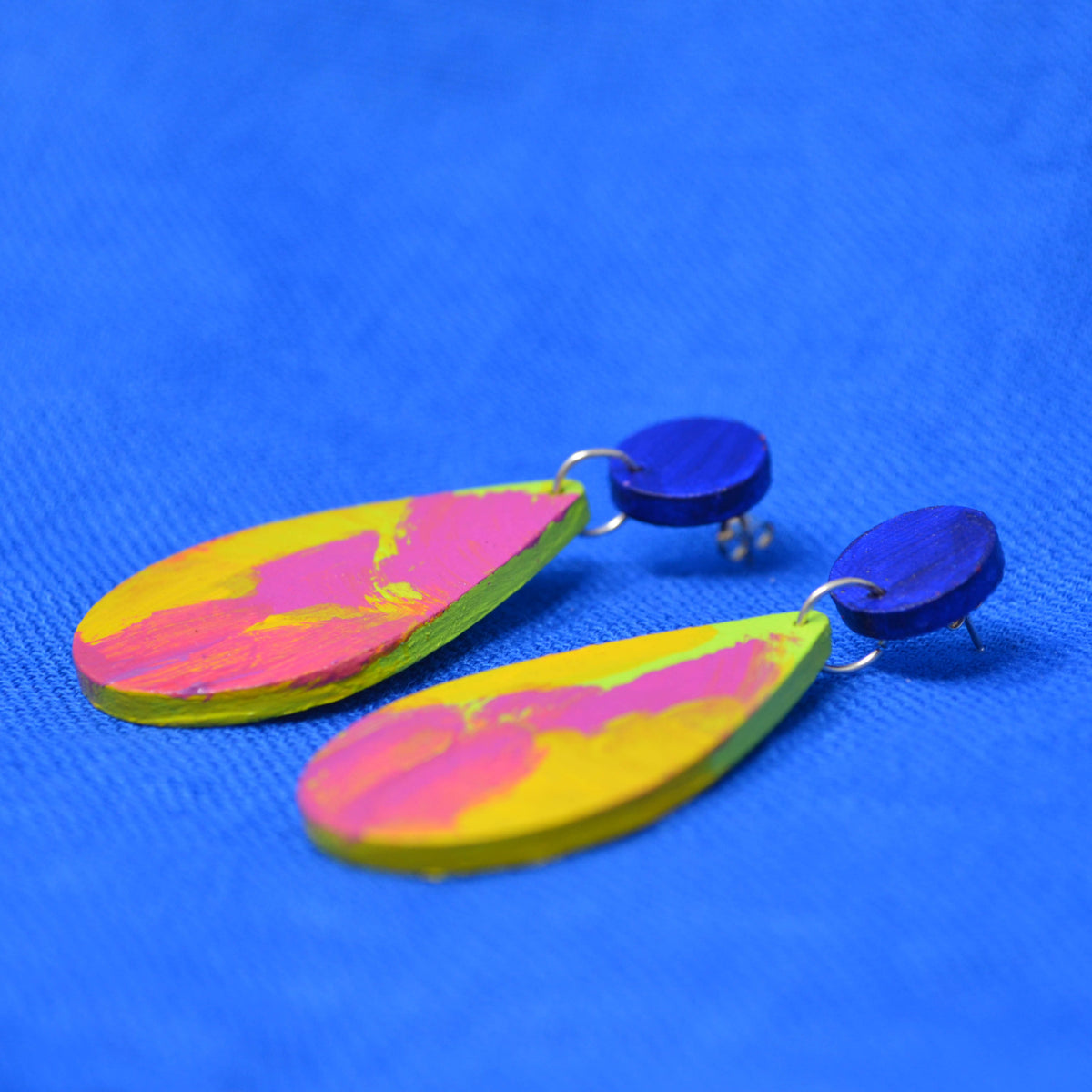 A pair of handmade drop earrings with dark blue circular studs and teardrop shaped dangles featuring a pink, green and yellow abstract painted design, displayed on a textured blue background.
