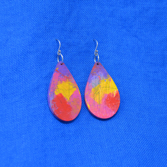 A pair of teardrop-shaped wooden dangle earrings with a colorful abstract paint design in red, orange, yellow, and purple, hanging from silver fishhook wires against a bright blue textured fabric background.