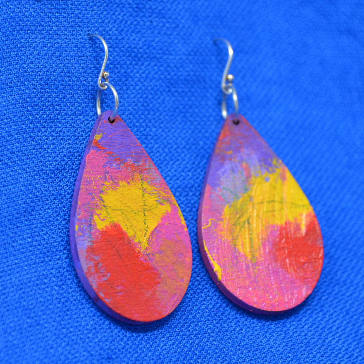 A pair of teardrop-shaped wooden dangle earrings with a colorful abstract paint design in red, yellow, pink and purple, hanging on silver fishhook wires against a bright blue textured fabric backdrop.