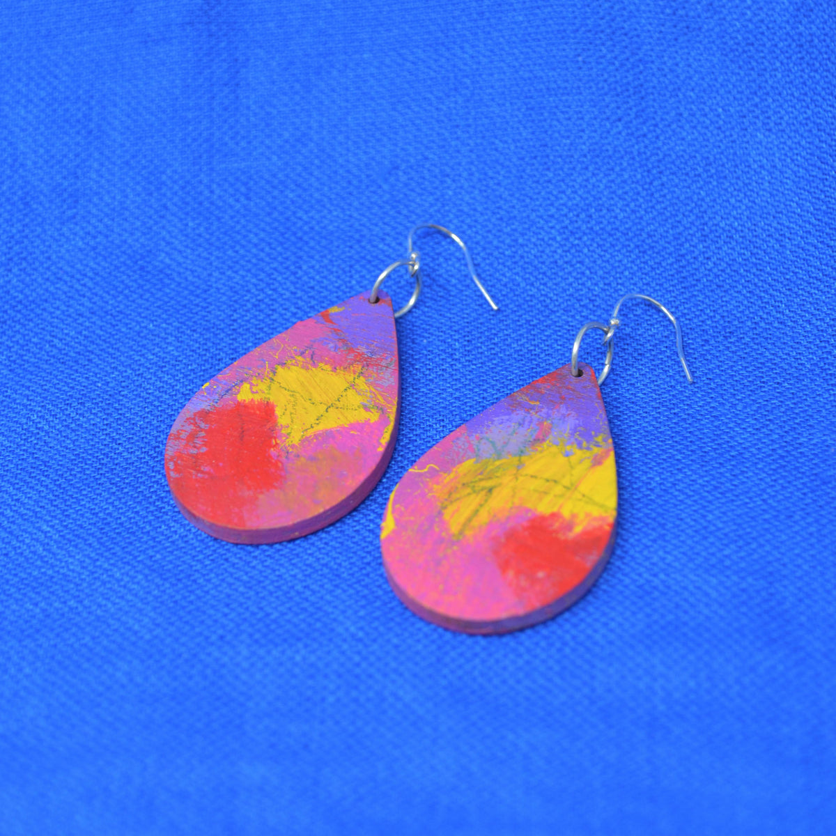 A pair of teardrop-shaped wooden earrings with a colorful, marbled pattern of pink, red, and yellow, featuring silver fishhook wires, displayed on a textured blue background.