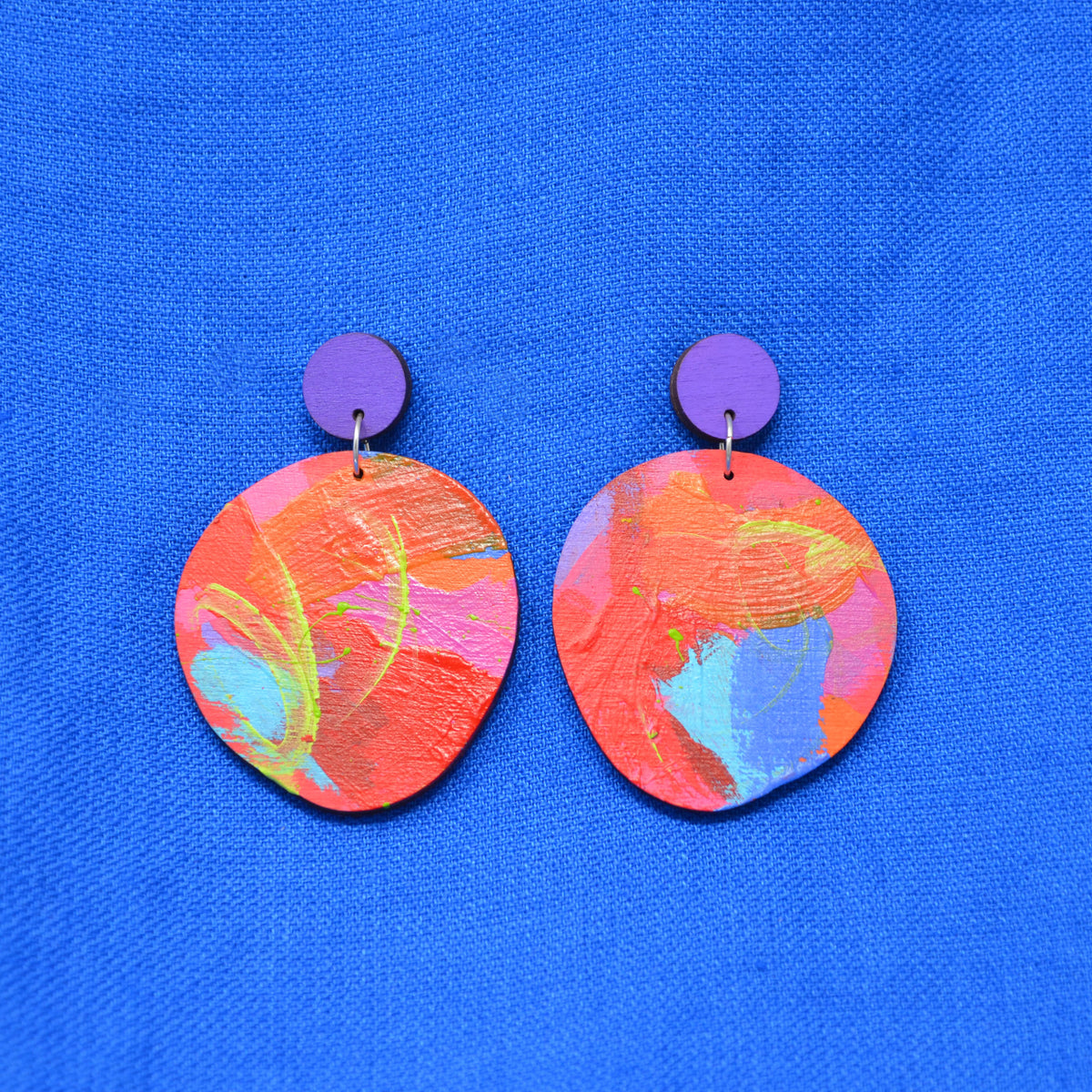 A pair of handmade drop earrings featuring circular orange, pink, and blue marbled painted discs hanging from solid purple circular studs, displayed on a textured blue fabric background.