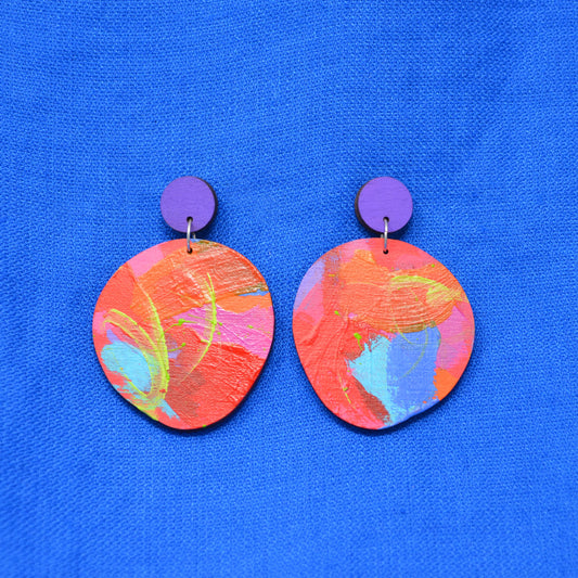 A pair of handmade drop earrings featuring circular orange, pink, and blue marbled painted discs hanging from solid purple circular studs, displayed on a textured blue fabric background.