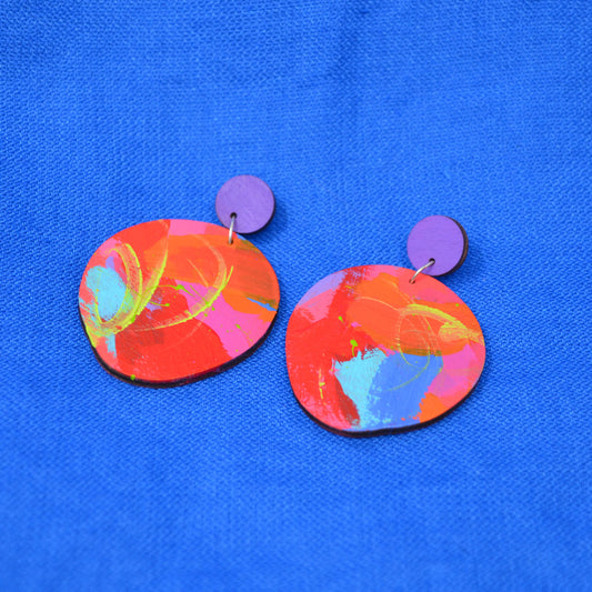 A pair of handmade earrings featuring abstract painted wooden discs in vibrant shades of red, orange, pink, and blue, with small purple circular studs, displayed on a textured blue background.
