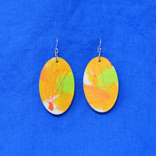 A pair of oval drop earrings with abstract painted designs in orange, yellow, green, and white, suspended from silver fishhook wires against a bright blue textured fabric background.