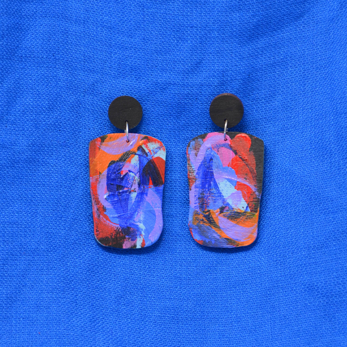 A pair of abstract painted rectangular drop earrings with black circular studs, featuring vibrant blue, red, orange, and purple brushstrokes, displayed on a textured blue fabric background.