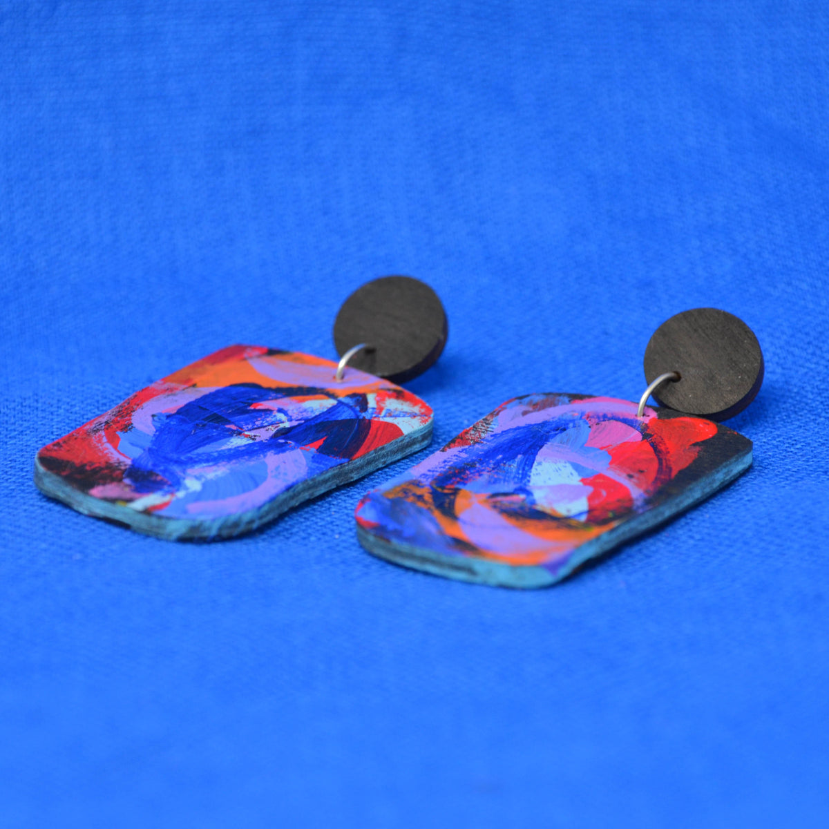 A pair of handmade rectangular drop earrings with colorful abstract painted wood tops and round black metal studs, displayed on a textured blue background.