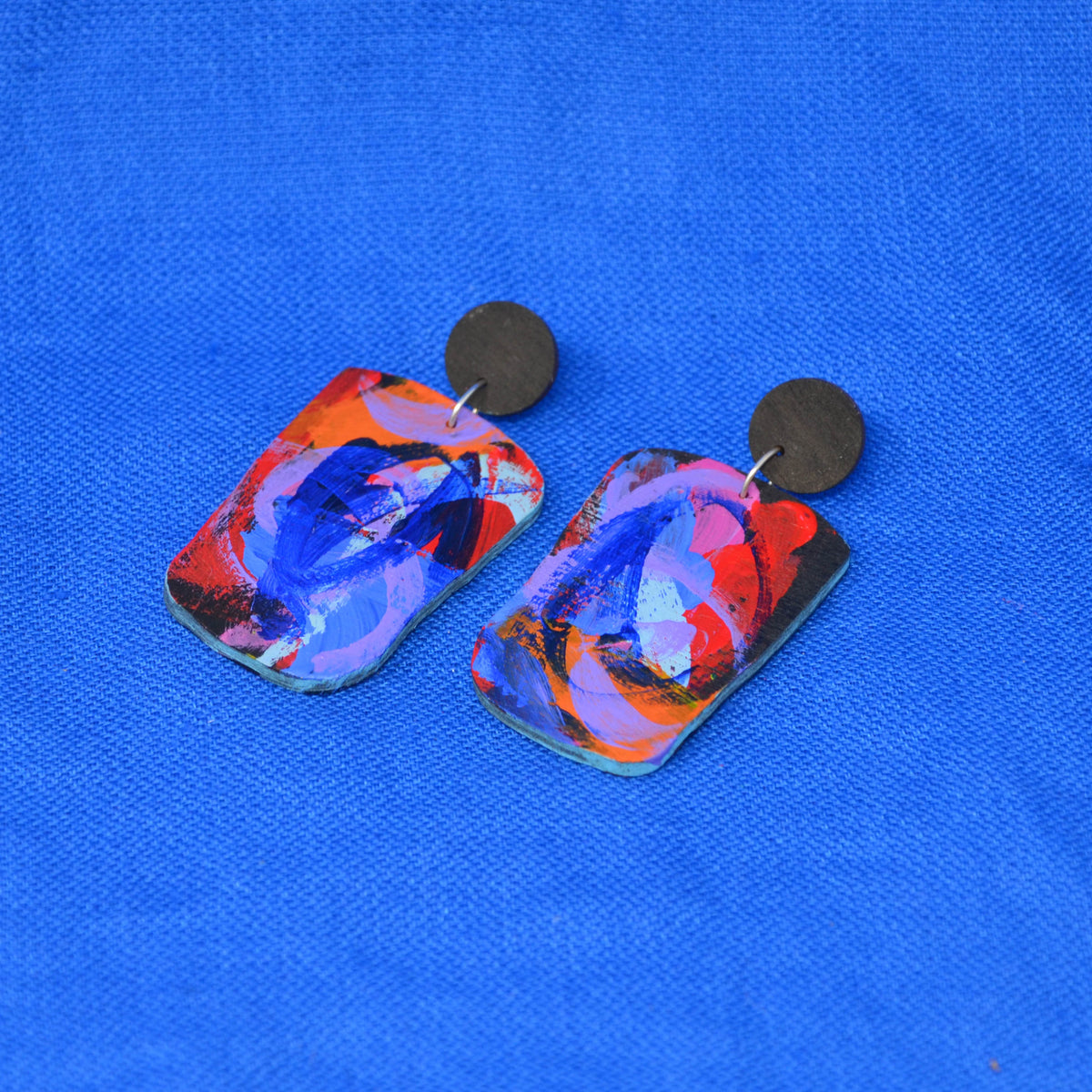 A pair of rectangular drop earrings with an abstract multicolor design in blue, red, orange, and black, hanging from dark round studs on a textured blue background.