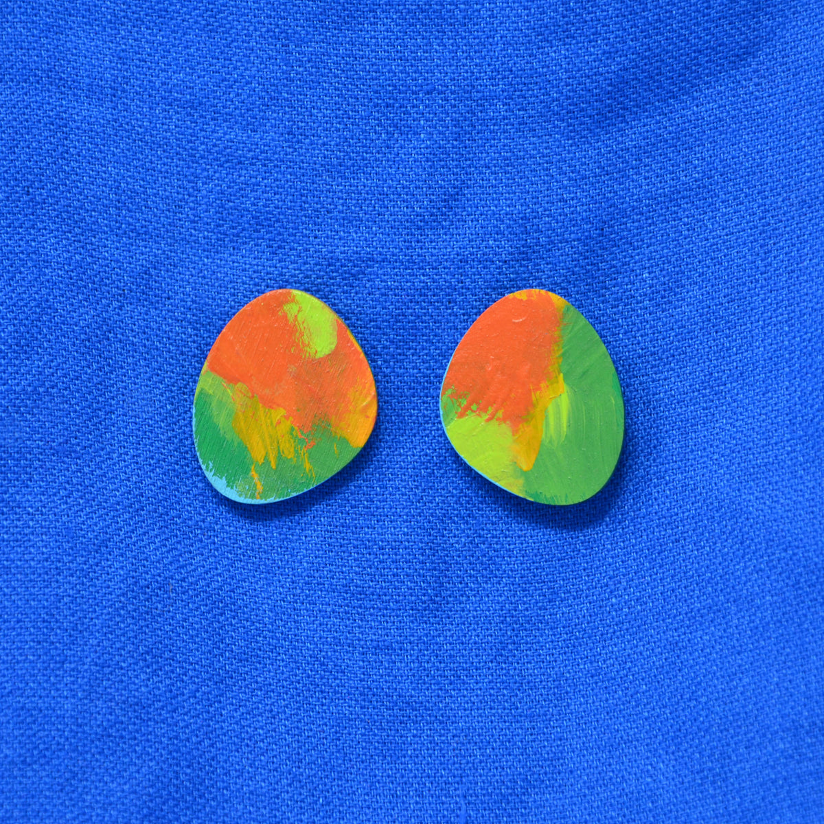 Pair of stud earrings with a colorful abstract paint design in orange, green, and yellow, displayed on a textured blue fabric background.