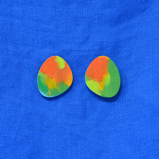 Pair of stud earrings with a colorful abstract paint design in orange, green, and yellow, displayed on a textured blue fabric background.