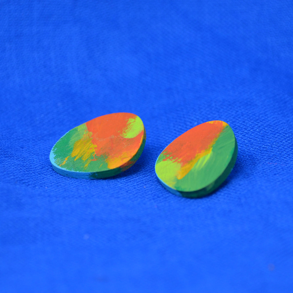A pair of teardrop-shaped wooden earrings featuring a vibrant, multicolored hand-painted design with swirls of orange, green, and yellow on a bright blue textured background.