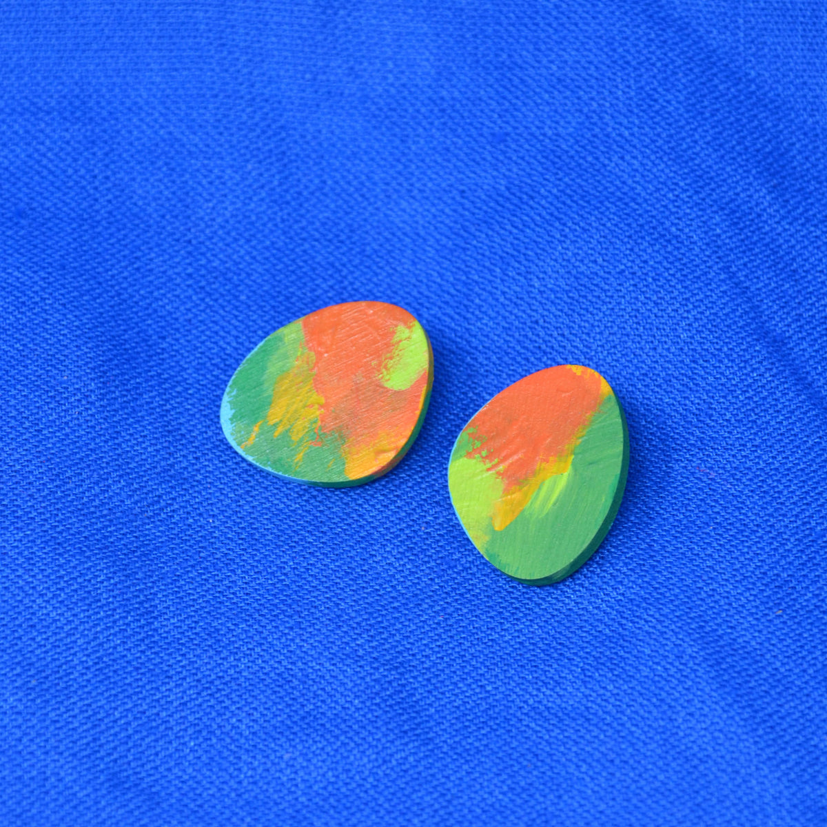 Pair of teardrop stud earrings painted with a marbled mix of green, orange, and yellow, placed on a bright blue textured fabric background.