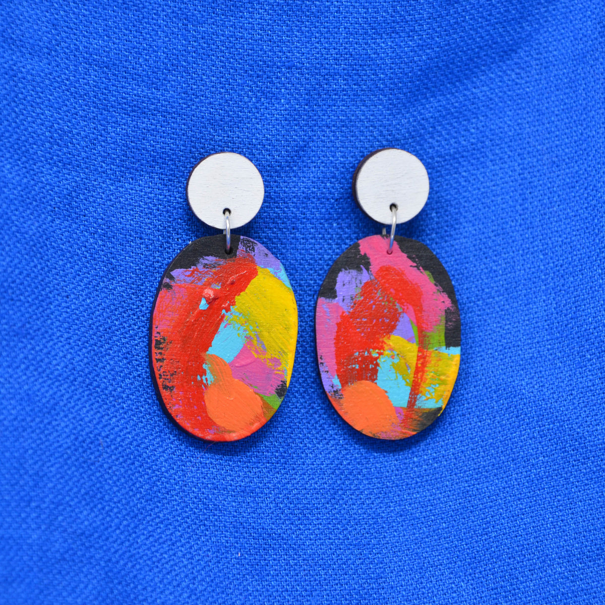 A pair of oval drop earrings with colorful abstract painted designs in red, orange, yellow, pink, and blue, hanging from white studs against a bright blue textured fabric background.