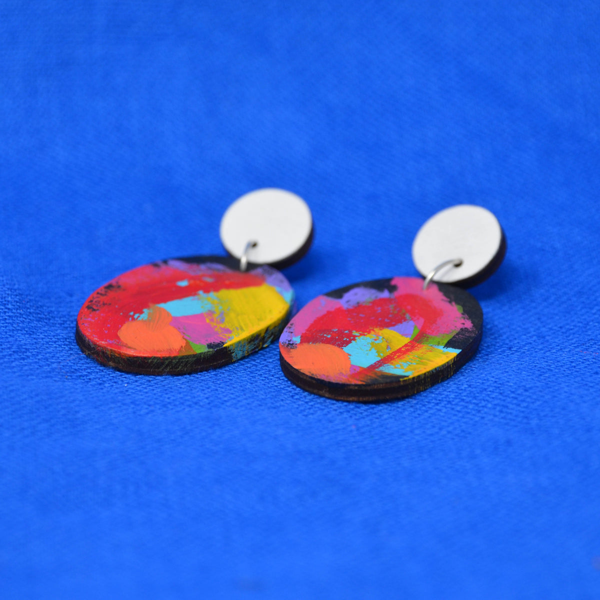 A pair of drop earrings with round white studs and oval wooden dangles featuring a vibrant, multicolored abstract paint splatter design, set against a textured blue background.