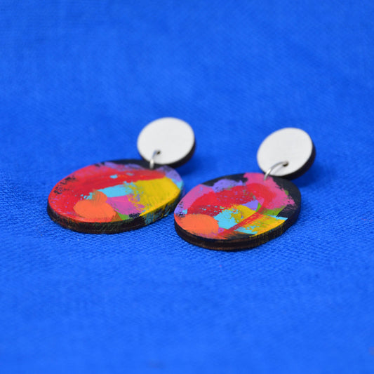 A pair of drop earrings with round white studs and oval wooden dangles featuring a vibrant, multicolored abstract paint splatter design, set against a textured blue background.
