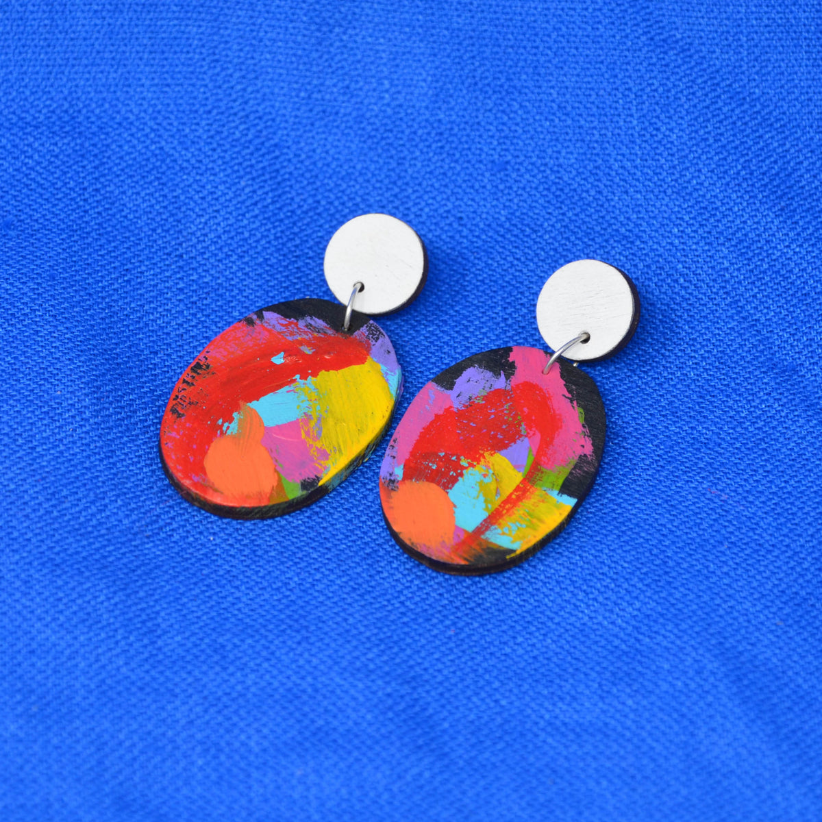 A pair of drop earrings with oval, abstractly painted charms in vibrant multicolor shades of red, yellow, pink, orange, and blue, suspended from round white discs, displayed on a textured blue background.