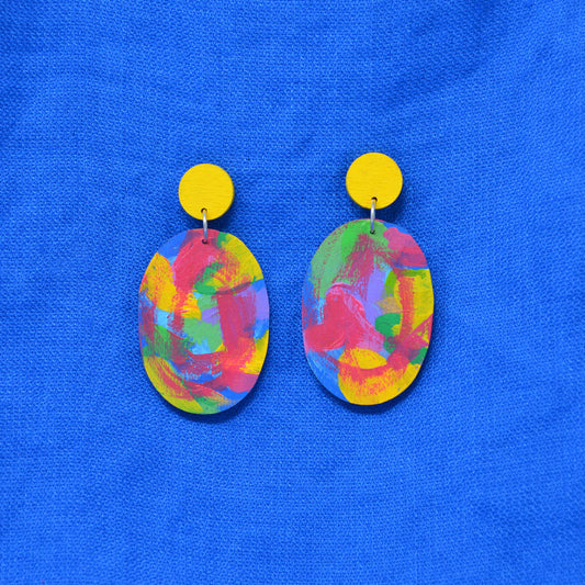 A pair of handmade drop earrings with bright yellow circular studs and oval charms featuring a colorful, abstract painted pattern in red, yellow, green, and blue, displayed on a textured blue fabric background.