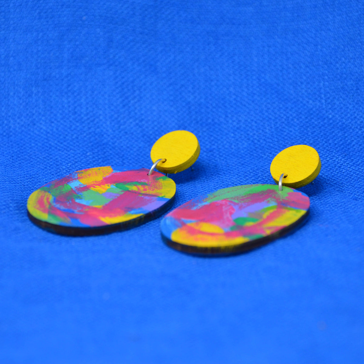 A pair of drop earrings with bright yellow circular studs and oval, multicolored painted wooden pendants, displayed on a textured blue background.