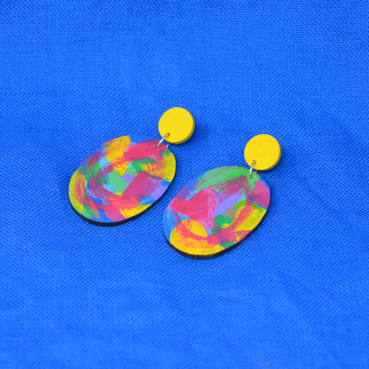 A pair of drop earrings with bright yellow circular studs and oval dangles featuring a colorful abstract paint-splatter design in pink, yellow, green, blue, and purple, displayed on a textured blue background.