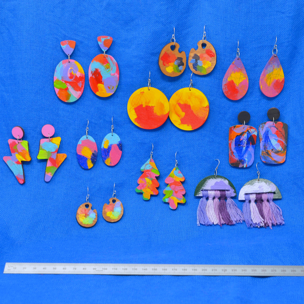 A collection of colorful handmade earrings arranged on a bright blue background, with a metal ruler at the bottom for scale. The earrings include abstract marbled and painted designs in red, orange, yellow, pink, blue, and green, plus some with purple tassels.