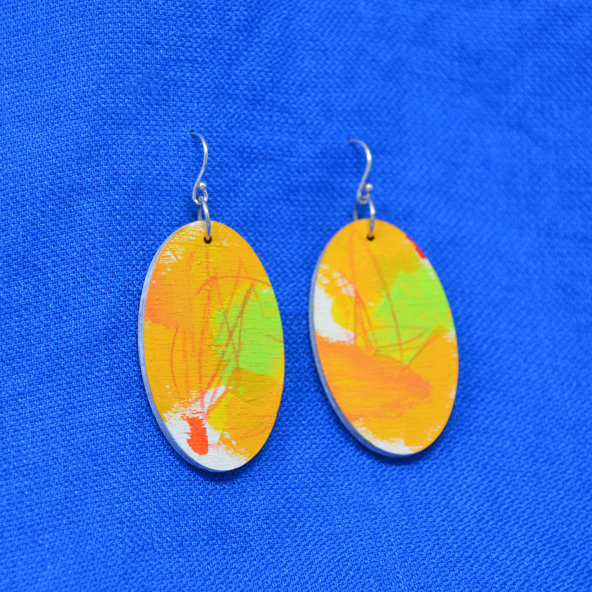 A pair of oval drop earrings with abstract painted designs in orange, yellow, green, and white, suspended from silver fishhook wires, displayed on a textured blue background.