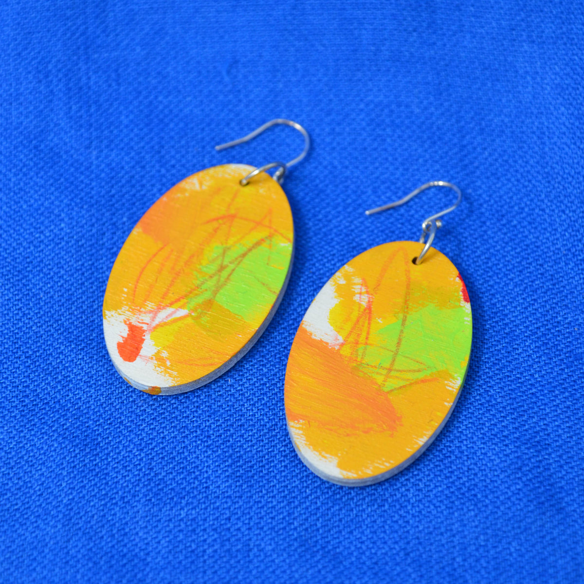 A pair of oval drop earrings with a colorful abstract paint design in orange, yellow, green, and white, hanging from silver fishhook wires on a textured blue background.