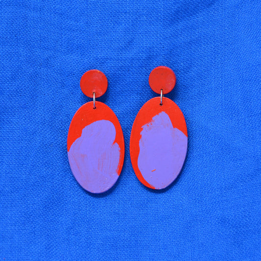 A pair of handmade drop earrings with red circular studs and teardrop shaped dangles featuring a red and purple abstract painted design, displayed on a textured blue background.
