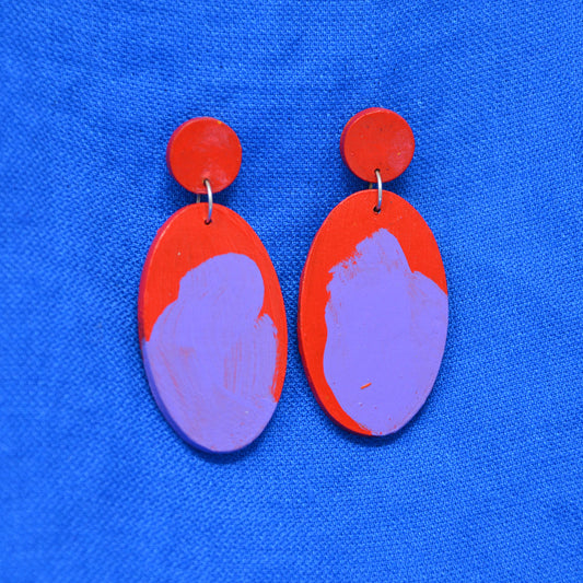 A pair of wooden handmade drop earrings with red circular studs and teardrop shaped dangles featuring a red and purple abstract painted design, displayed on a textured blue background.