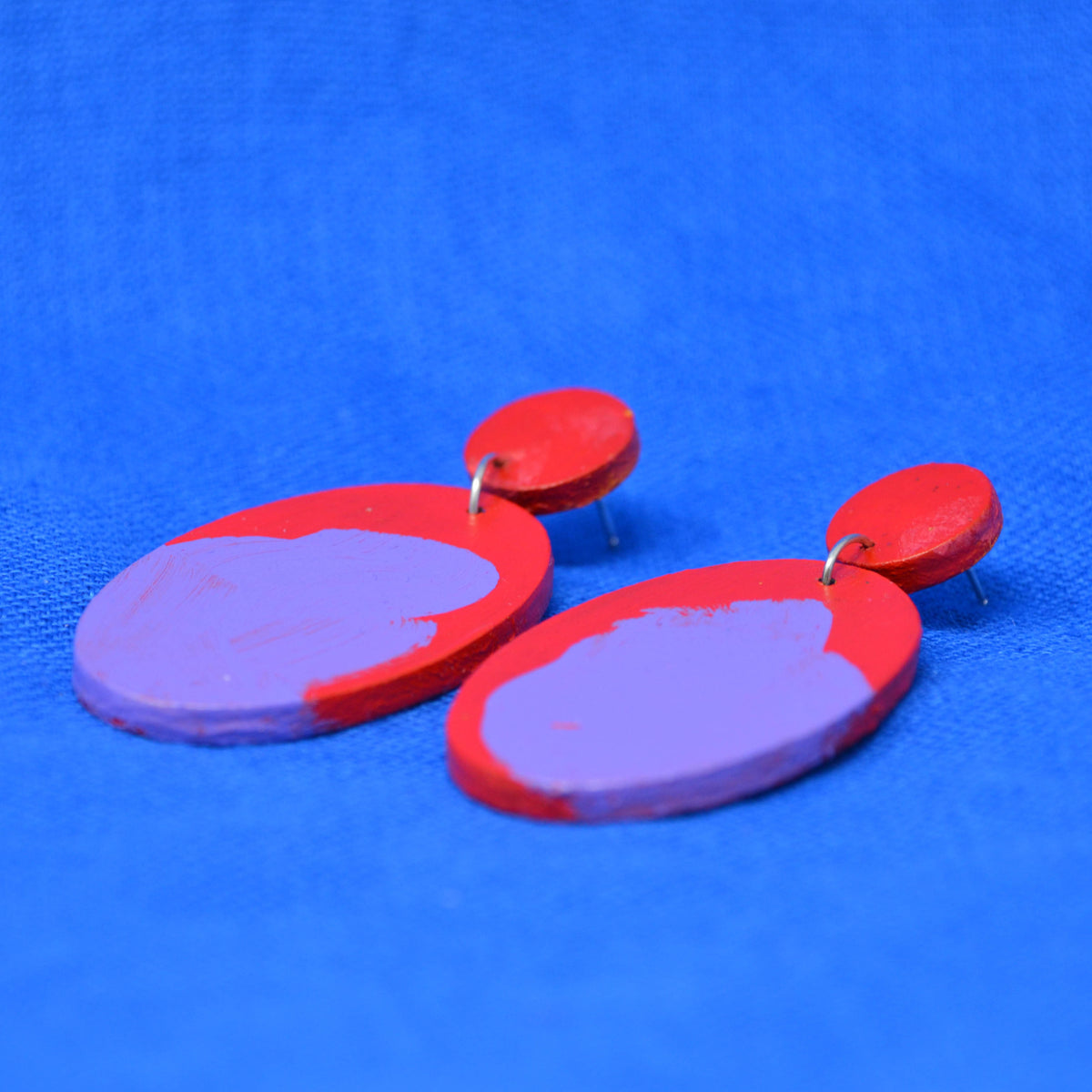 A pair of handmade drop earrings with red circular studs and teardrop shaped dangles featuring a red and purple abstract painted design, displayed on a textured blue background.