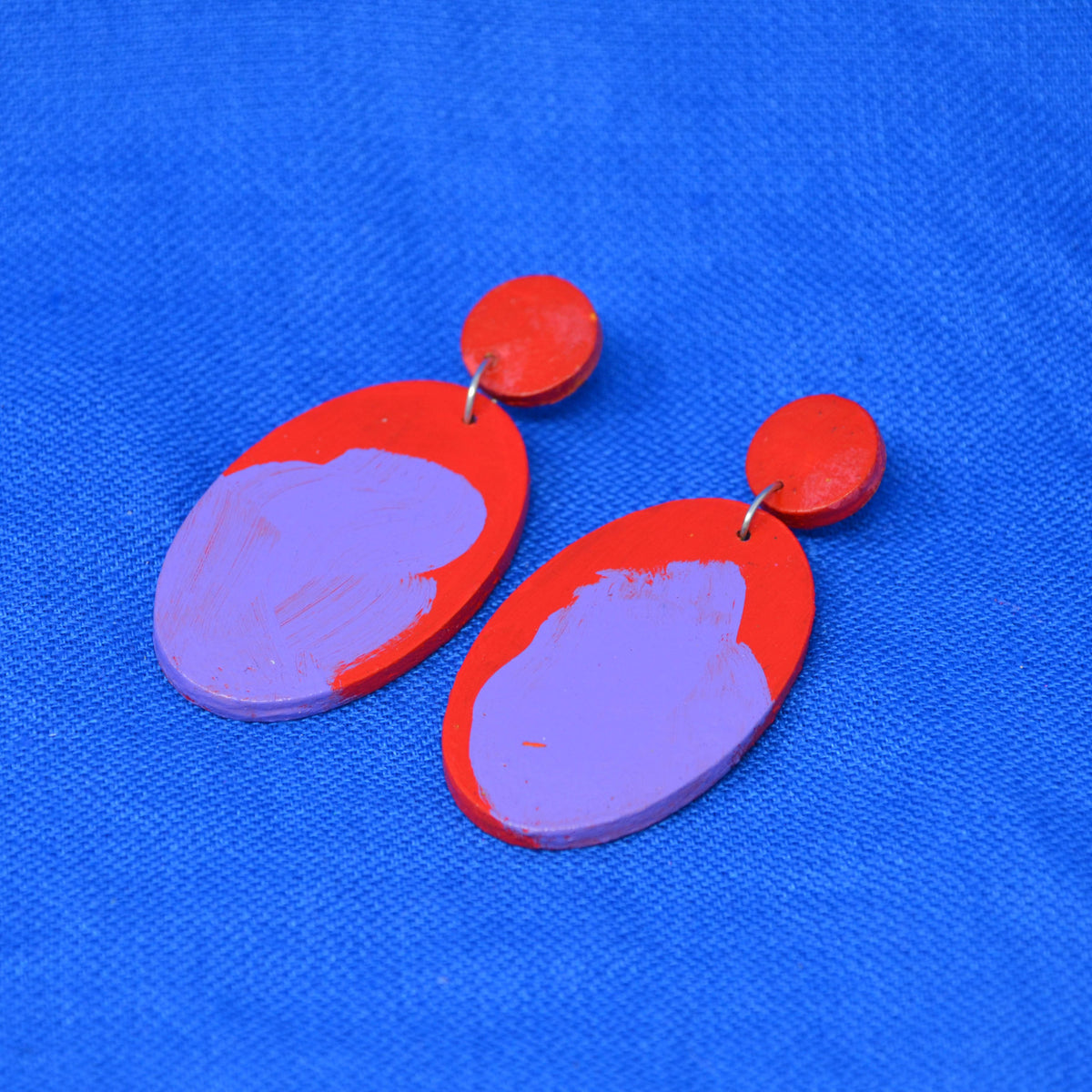 A pair of handmade drop earrings with red circular studs and teardrop shaped dangles featuring a red and purple abstract painted design, displayed on a textured blue background.